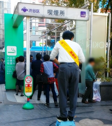 渋谷駅前のハチ公横の喫煙所の前に立つ、コミヤマさん。